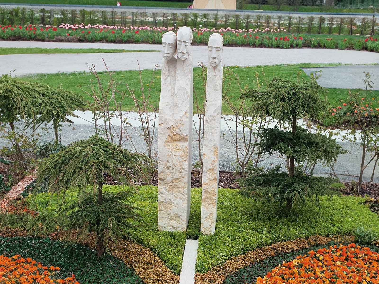 Grabstein aus drei Säulen, die jeweils eien Person symbolisieren. Es sind grob behandelte Steine mit oben eingearbeiteten Köpfen.